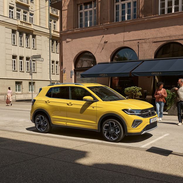 Ein gelber VW T-Cross parkt am Straßenrand in einer Stadt mit mehrstöckigen Gebäuden, ein Pärchen geht darauf zu.