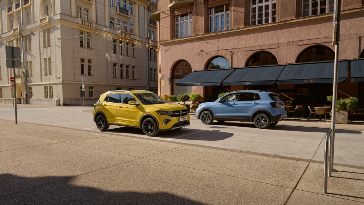 Ein gelber VW T-Cross parkt am Straßenrand in einer Stadt. Ein blauer T-Cross fährt daran vorbei.