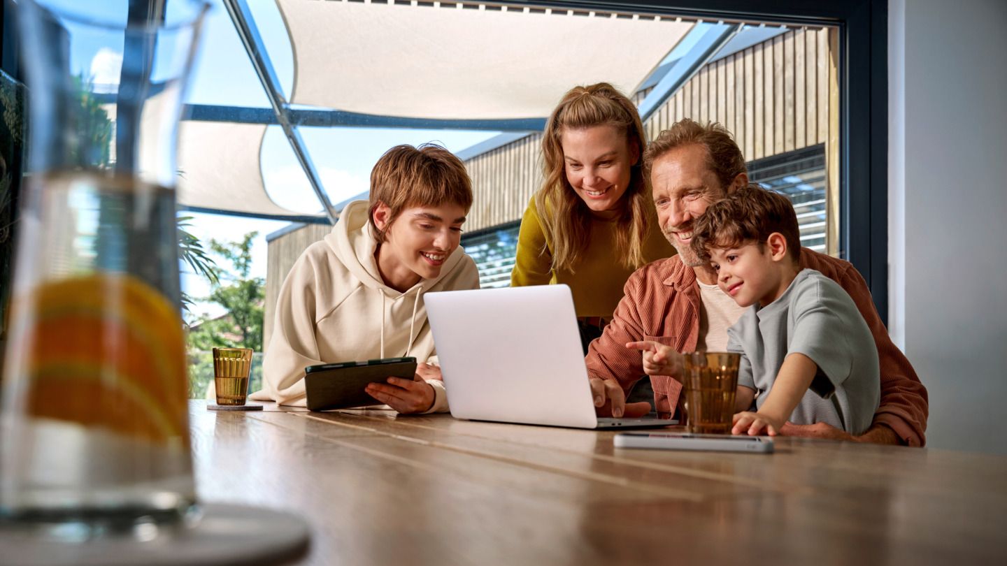 Eine Familie ist um einen Tisch versammelt und schaut fröhlich auf einen Laptop-Bildschirm