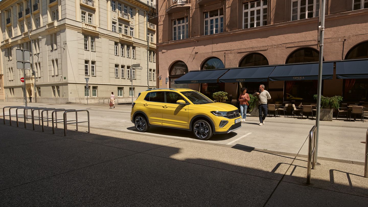 Ein gelber VW T-Cross parkt am Straßenrand in einer Stadt mit mehrstöckigen Gebäuden, ein Pärchen geht darauf zu.