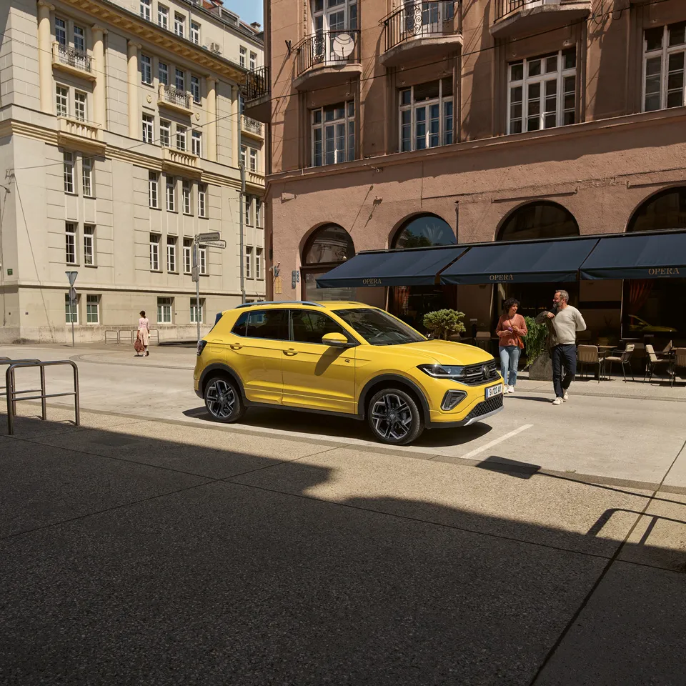 Ein gelber VW T-Cross parkt am Straßenrand in einer Stadt mit mehrstöckigen Gebäuden, ein Pärchen geht darauf zu.