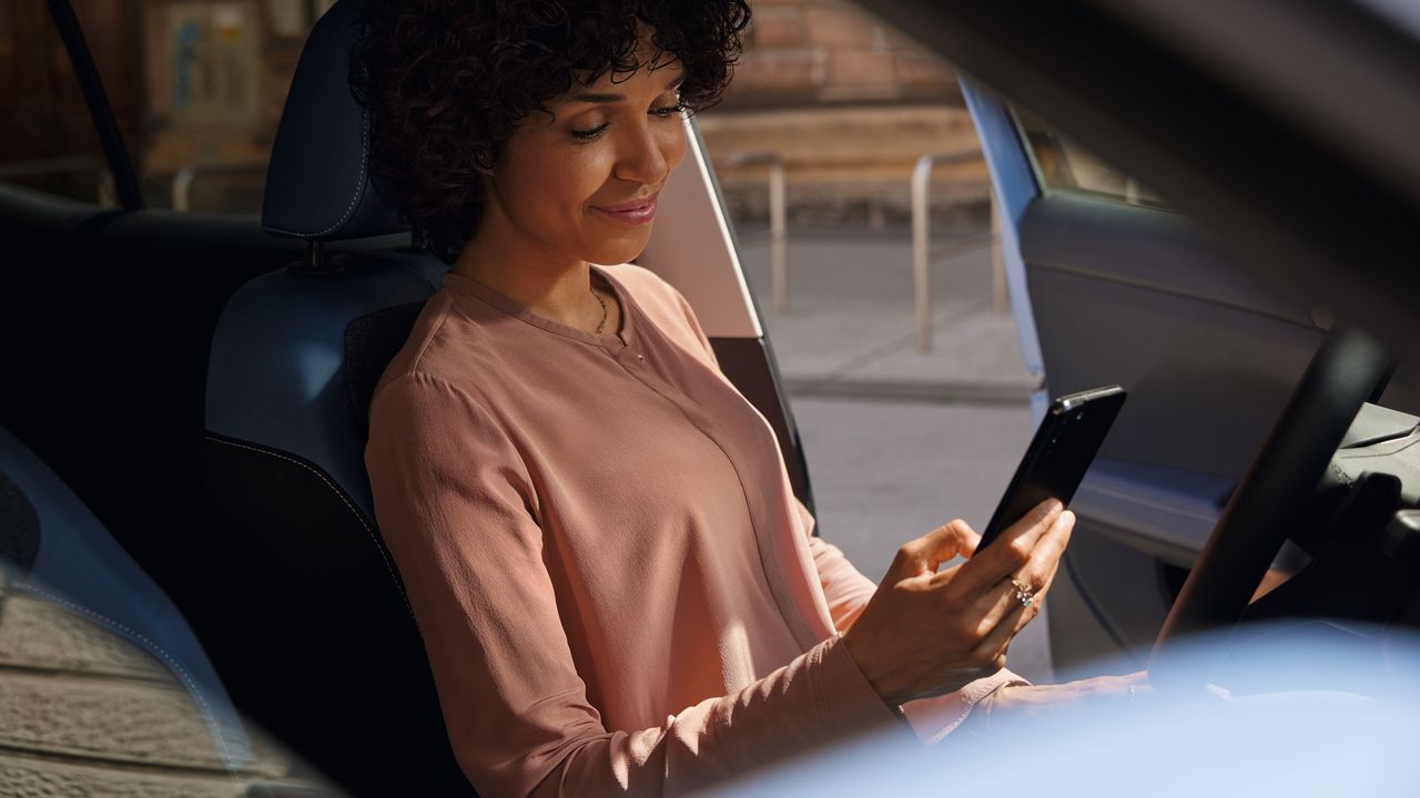 Eine lächelnde Frau sitzt auf dem Fahrersitz eines VW T-Cross und schaut auf ihr Smartphone.