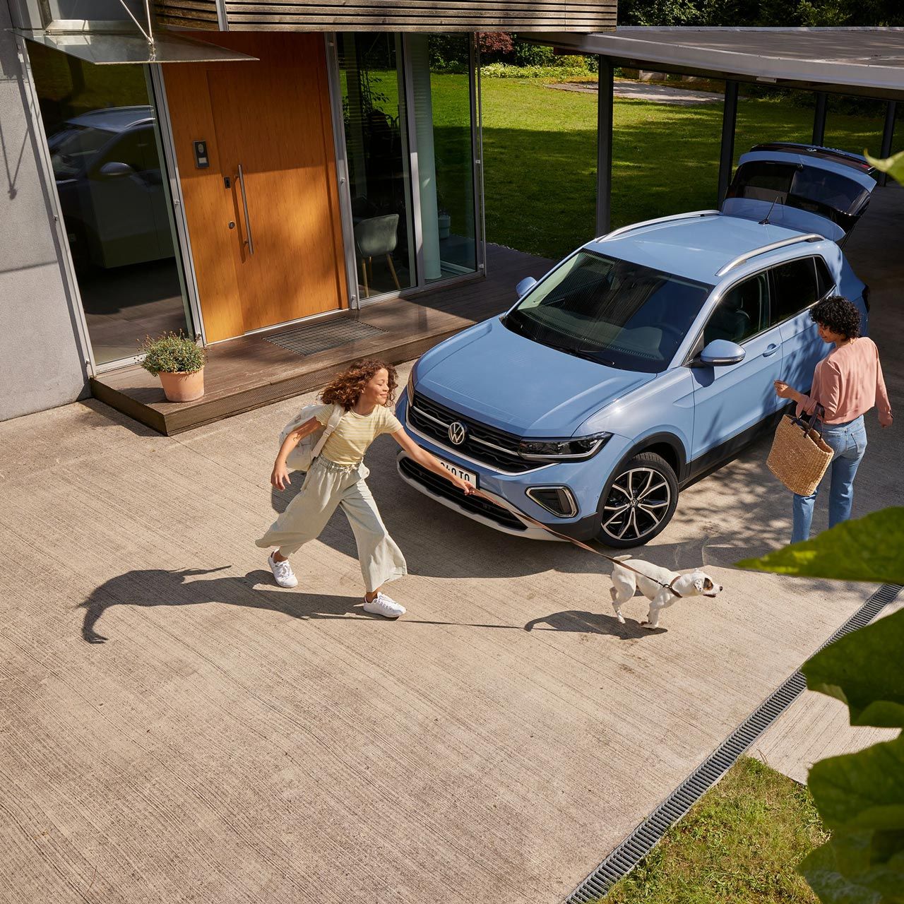 Eine Frau mit Korb und ein Mädchen mit Hund vor einem blauen VW T-Cross mit geöffnetem Kofferraum in einer Einfahrt.