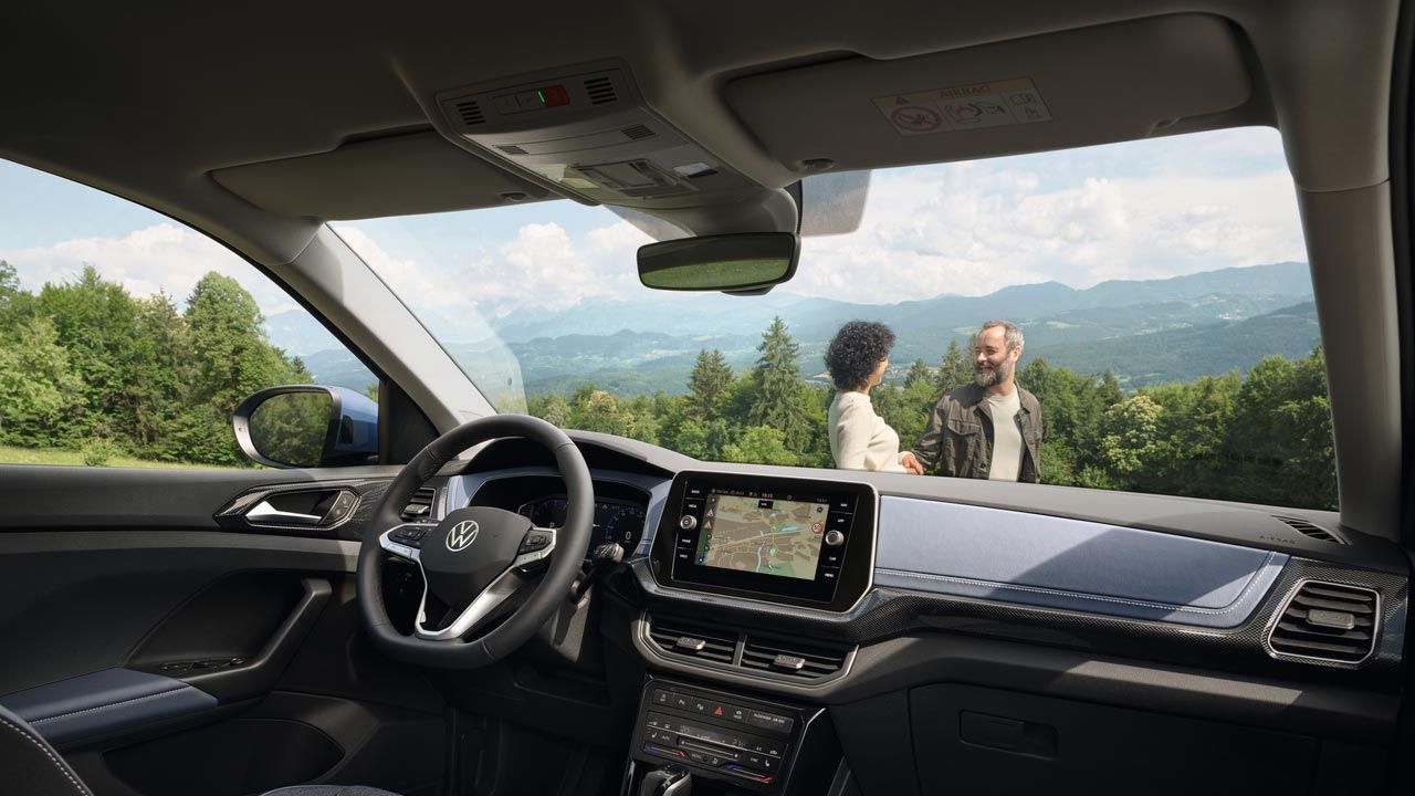 Blick vom Beifahrersitz eines VW T-Cross durch die Frontscheibe auf ein lachendes Pärchen vor Wald und Bergen.