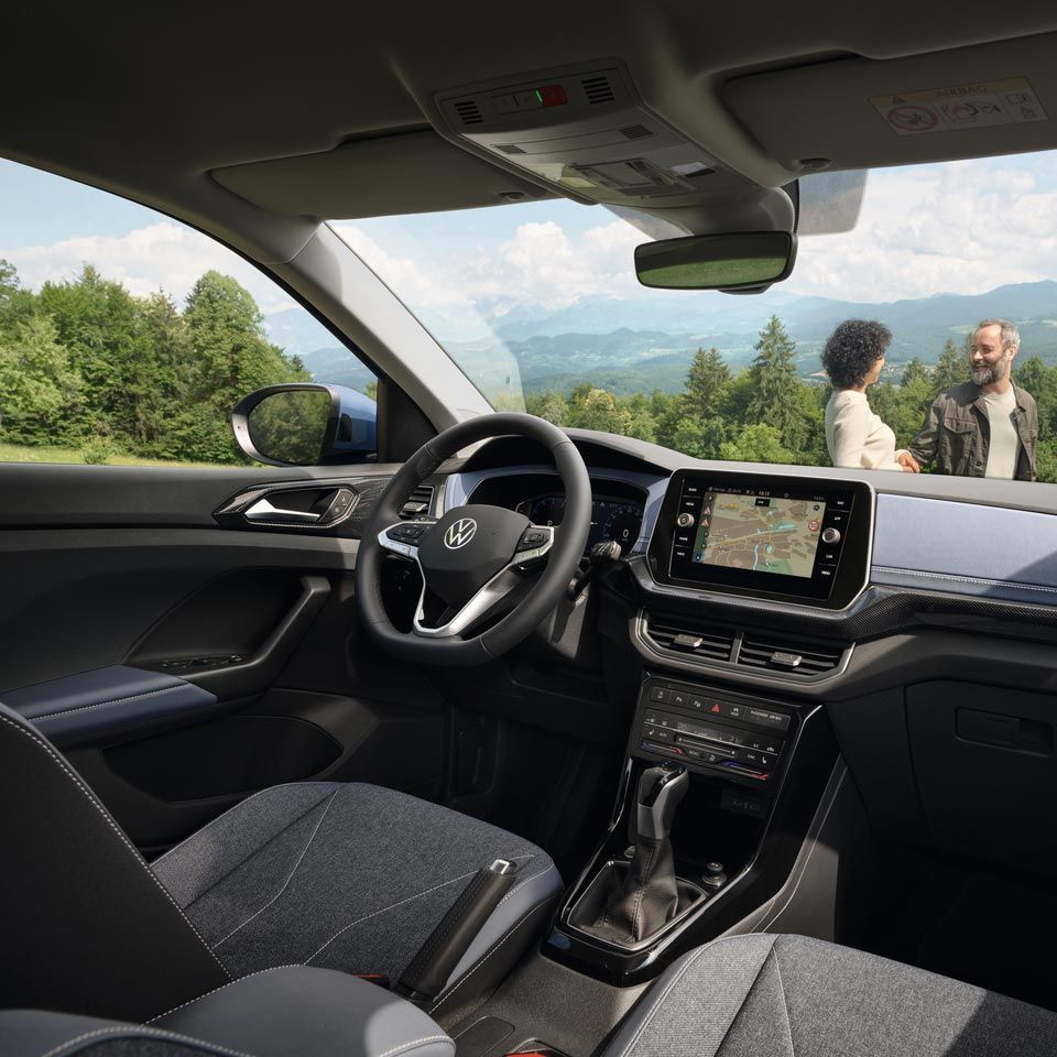 Blick vom Beifahrersitz eines VW T-Cross durch die Frontscheibe auf ein lachendes Pärchen vor Wald und Bergen.