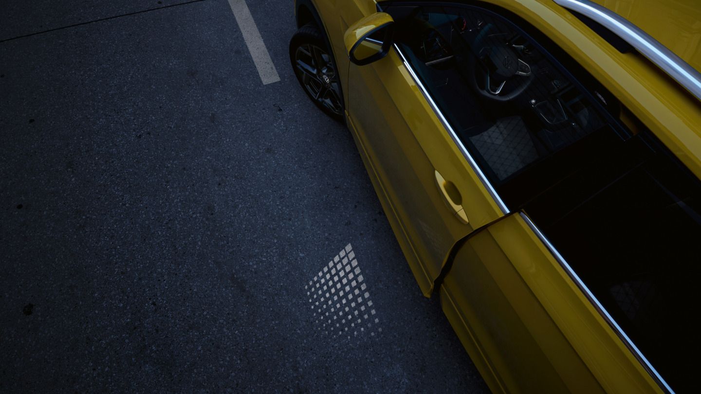 Die leicht geöffnete Fahrertür eines gelben VW T-Cross lässt die Lichtprojektion auf den Parkplatzboden scheinen.